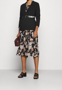 Schwarze taillierte Blazerjacke mit langen Ärmeln und einem Gürtel, über einem schwarzen Blumenkleid. Die Person trägt Sandalen mit Blockabsätzen und hält eine kastanienbraune Tasche.