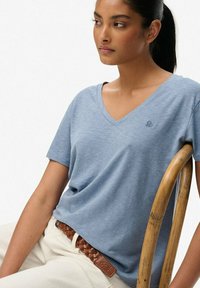Blå v-hals t-shirt lavet af et blødt, struktureret stof. Har korte ærmer og et subtilt logo. Bæres med hvide bukser og et flettet brunt bælte.