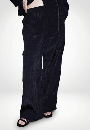 Persona vistiendo pantalones negros sueltos con textura y cinturilla elástica, y sandalias negras abiertas en los dedos, de pie con una mano en la cadera.