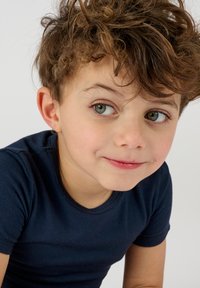 Bambino con capelli ricci castani, che indossa una maglietta a maniche corte blu navy. Ha la pelle chiara e occhi verde-blu. Appare pensieroso.