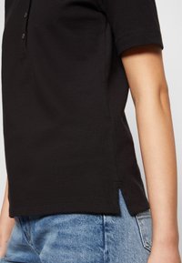 Schwarzes Polohemd mit kurzen Ärmeln und klassischem Kragen, mit einer seitlichen Schlitz-Detailierung und strukturiertem Stoff. Kombiniert mit hellblauen Jeans.