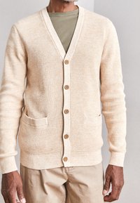 Beige knitted cardigan with a V-neck, wooden buttons, two front pockets, and ribbed cuffs. Layered over a green T-shirt.