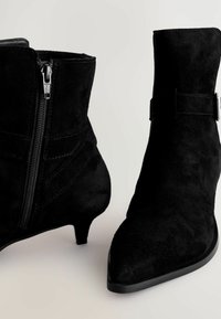 Des bottines en suede noir présentent un bout pointu, un petit talon, une fermeture éclair sur le côté et un détail de sangle avec des accents métalliques.