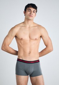 Grå boksertruser med en svart og rød HUGO-merket linning, laget av et glatt og tettsittende materiale. Modellen står med avslappede armer.