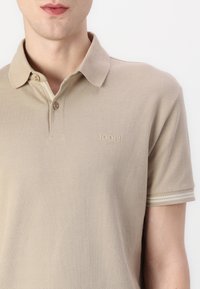 Bežinis tekstūruotas polo marškinėliai su apykakle, dviejų mygtukų užsegimu ir dryžuotais akcentais ant rankovių; krūtinėje siuvinėti logotipai.