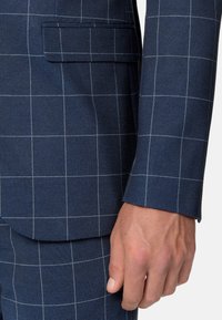 Chaqueta de traje azul marino con un patrón de cuadrícula. Cuenta con un bolsillo y un corte entallado. La tela presenta una textura sutil y costuras en contraste.