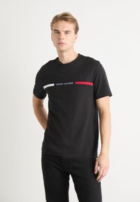 Schwarzes Baumwoll-T-Shirt mit Rundhalsausschnitt. Verfügt über ein horizontales Grafikdesign in Weiß, Rot und Blau mit dem Schriftzug "TOMMY HILFIGER" in der Mitte.