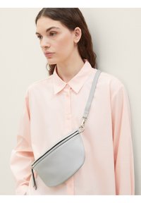 Gray leather crossbody bag with a curved shape, single zip closure, and adjustable strap, worn over a pale pink button-up shirt.