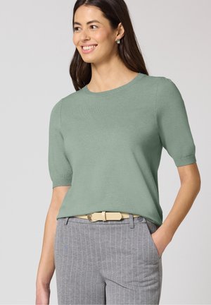 Femme souriante portant un pull léger à manches courtes vert clair, rentré dans un pantalon gris à fines rayures avec une ceinture beige, main dans la poche, posant devant un fond uni.