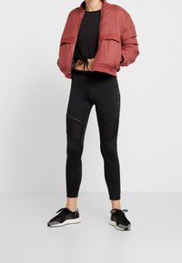 Czerwona, pikowana kurtka bomberka, czarny top crop, czarne legginsy z siateczkowymi detalami oraz czarne buty sportowe z białymi akcentami.