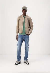 Beige bomberjakke med ribbestrikkede mansjetter, grønn t-skjorte, lyseblå jeans og sølv sneakers. Caps med brodert tekst fullfører antrekket.