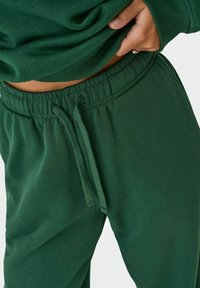 Grønne sweatpants med elastisk buksering, snor-detail og en glat tekstur, delvist synlige under en matchende top.