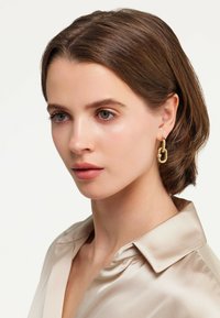 Boucles d'oreilles en métal doré avec un design en chaîne liée et un petit accent en perle. Portées par un mannequin ayant des cheveux bruns mi-longs, dans une chemise en satin.