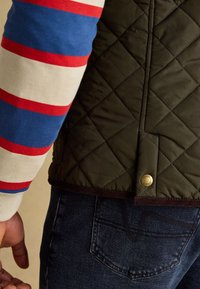 Nahaufnahme einer Person, die eine gesteppte dunkelgrüne Jacke, blaue Jeans und ein gestreiftes langärmeliges Hemd mit roten, blauen und weißen Streifen trägt.
