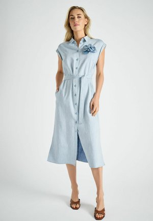 Vrouw staand tegen een effen achtergrond, gekleed in een lichtblauwe midi-jurk met knopen, tailleceintuur, bloemenbroche en bruine openhakken.