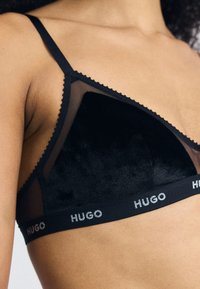 Black bra featuring sheer mesh and soft velvet materials, triangular cups, scalloped edges, and a branded elastic band with "HUGO."