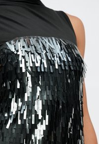 Μαύρο αμάνικο τοπ με λαμπερά κρόσσια από αντανακλαστικές λωρίδες, δημιουργώντας μια στρωματοποιημένη υφή στην περιοχή του στήθους.