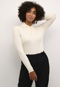 Polo a maniche lunghe color crema con texture a coste e zip frontale, abbinato a pantaloni neri a vita alta.