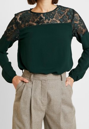 Donna che indossa una blusa verde scuro a maniche lunghe con spalla in pizzo floreale e pantaloni beige plissettati a vita alta, con le mani nelle tasche.