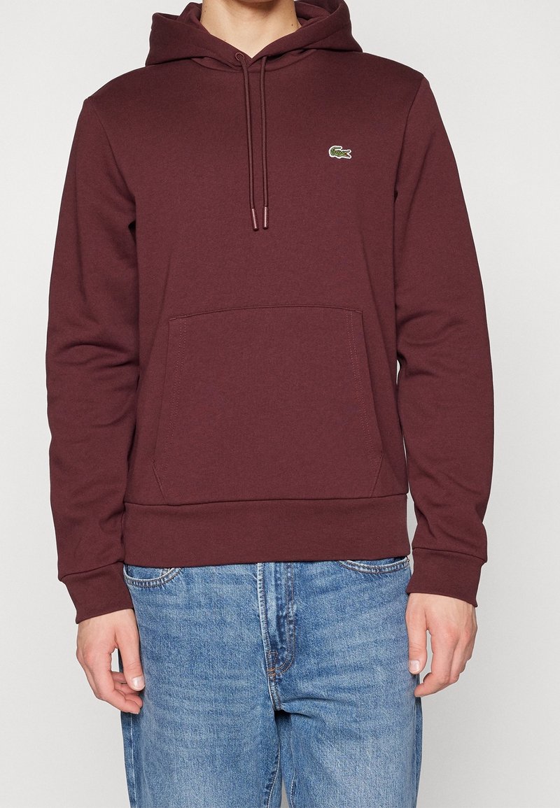 Homme portant un sweat à capuche bordeaux avec une poche avant et un petit logo crocodile vert, associé à un jean bleu.