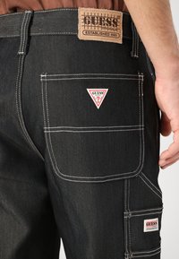 Sorte denimbukser med hvite sømmer, to sidelommer og Guess-logo lapper på liv og lommer. Holdbart, teksturert stoff.