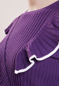 Haut violet côtelé avec un col rond et des accents de volants asymétriques. Les volants sont bordés de détails blancs pour un contraste.