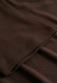 Rjava bombažna tkanina z gladko teksturo, ki prikazuje subtilen rebrast vzorec in natančno izšite robove. Gube ustvarjajo mehke drape.