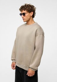 Beige sweatshirt met een oversized pasvorm, ronde halslijn en geribbelde manchetten, gecombineerd met donkere broeken en een zonnebril. Vriendelijke textuur, minimalistisch ontwerp.