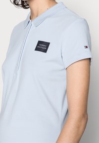 Světle modré polokošile vyrobené z bavlny, se černým čtvercovým logem s nápisem "TOMMY HILFIGER". Krátké rukávy s malým akcentem značky na manžetě.