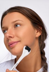 Vit dermaroller hålls mot en kind, med ett handtag med texturerat grepp och fina svarta nålar. Mjuk hud med neutrala toner.