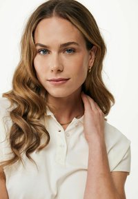 Ljus krämfärgad polo med korta ärmar, med en placket med tre knappar och textil med struktur. Modellen bär ett guldhalsband och örhängen.