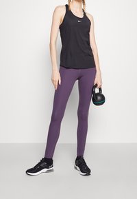 Zwarte Nike tanktop, paarse leggings en zwarte sneakers met witte accenten. De persoon houdt een zwarte kettlebell vast met een blauwe streep.