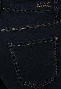 Donkerblauwe denim jeans met een achterzak. Heeft zichtbare stiksels in lichtbruin en een geborduurd logo in dezelfde kleur nabij de tailleband.