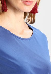Niebieska bluzka z krótkim rękawem i okrągłym dekoltem, wykonana z gładkiego materiału. Czerwone kolczyki z frędzlami dodają kontrastu. Skupienie na okolicy szyi i ramion.