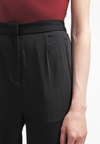 Pantalon noir sur mesure avec une texture lisse, une taille haute et des plis devant. Un bras repose sur la taille, mettant en valeur la coupe du vêtement.