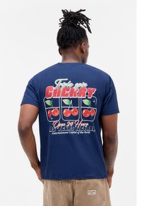 Man with dreadlocks wearing navy t-shirt with "Triple spin cherry" and Las Vegas slot machine graphic on back, paired with beige shorts.
