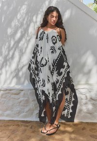 Črno-bela kaftan s potiskom in lahkotnim dizajnom, z odprtim sprednjim delom, trakovi za ramena in teksturirano tkanino. Nosi se z sandalmi.