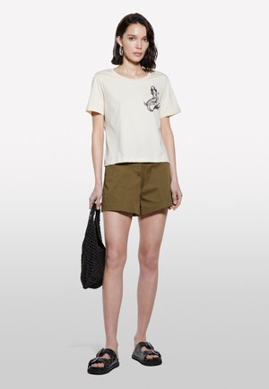 Cremfarbendes T-Shirt mit kurzen Ärmeln und einem Meerjungfrauen-Motiv, kombiniert mit olivgrünen Shorts. Eine schwarze geflochtene Tasche und klobige schwarze Sandalen runden den Look ab.