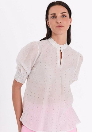 Vrouw met donker haar draagt een witte, gestructureerde kortermige blouse met een gerimpelde kraag en sleutelgathals, gecombineerd met lichtroze broek.