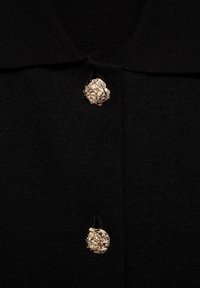 Vêtement en tricot noir avec un col et deux boutons décoratifs dorés ornés d'un motif floral complexe.