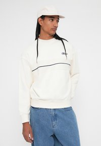 Svetlo krémový sveter s námorníckou pruhou, ktorý má logo na ľavej hrudi. Model má na sebe svetlo modré džínsy a zodpovedajúcu čiapku.