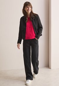 Giacca nera con zip, blusa rossa in tessuto strutturato con bottoni, pantaloni neri a gamba larga e sneakers bianche. Materiali morbidi e design casual.