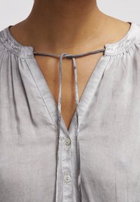 Blouse gris clair avec un col en V, dotée d'un détail noué au niveau du col, de plis délicats et d'une fermeture à boutons. Texture lisse, design décontracté.