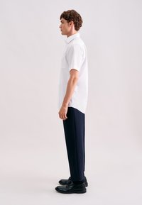 Wit overhemd met korte mouwen en knopen, voorzien van een zak, gecombineerd met marineblauwe broek en zwarte schoenen, van opzij weergegeven tegen een neutrale achtergrond.