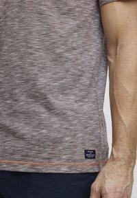 Brown and gray striped short-sleeve t-shirt with textured fabric, featuring a visible tag at the hem and contrasting orange stitching.