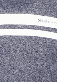 Tkany materiał w kolorze granatowym z melanzem, z dwoma poziomymi białymi pasami, z jednym z nich zawierającym częściowy tekst "DENIM TOM TAI" na odzieży.