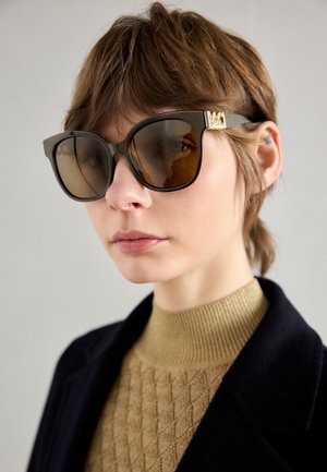 Bruine oversized zonnebril met dikke monturen en gouden accenten, met een gladde textuur en een unieke cat-eye vorm.