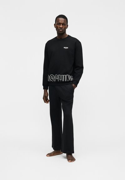 Άνδρας όρθιος, ξυπόλυτος, ντυμένος με μαύρο φούτερ Moschino και μαύρο παντελόνι, μπροστά σε λευκό φόντο.