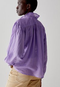Camicia viola a maniche lunghe con colletto alto, dalla vestibilità ampia e leggermente trasparente. Indossata con pantaloni chiari.