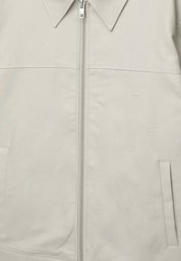 Chaqueta gris claro con cremallera frontal, cuello ancho y dos bolsillos laterales. Textura lisa y diseño sencillo sin patrones ni detalles visibles.